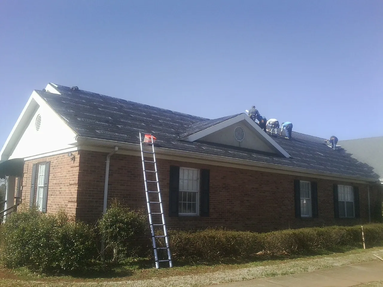 Crew tearing shingles off a brick ranch roof with a ladder set — Metal Roof Repair work by our crew in Hanscom Afb