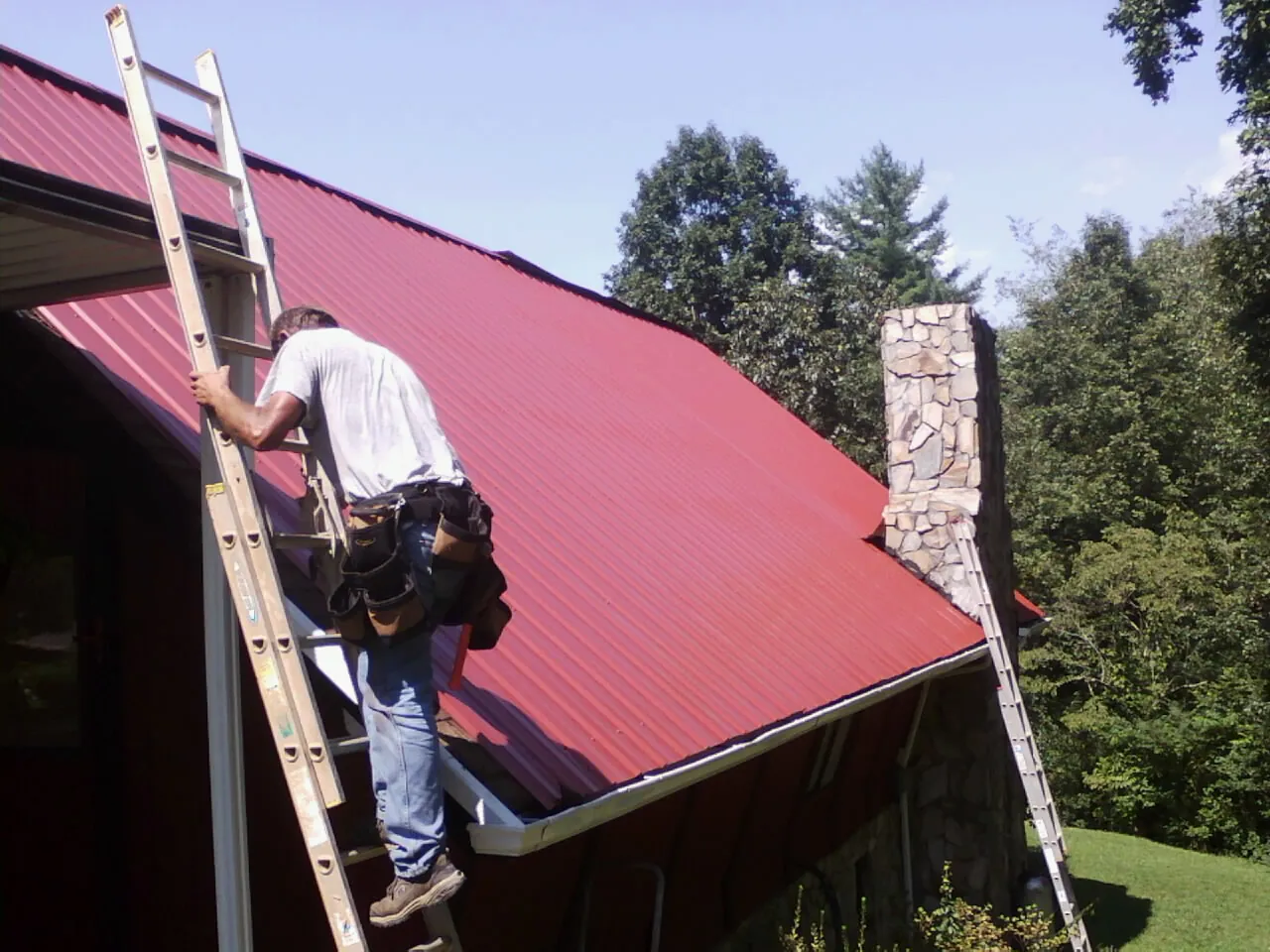 Roofer on a ladder beside a red metal roof and stone chimney — Soffit & Fascia Repair by Roofing Brothers in Hanscom Afb