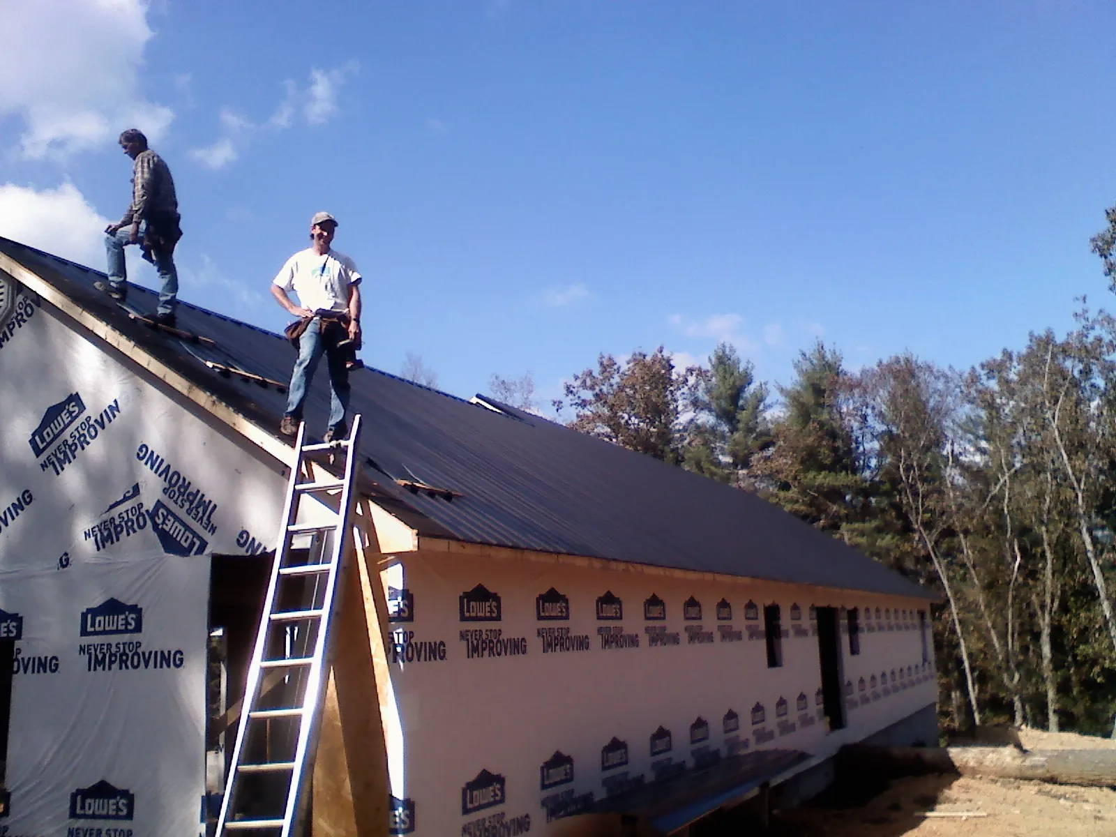 Two roofers standing on a long dark metal roof over a wrapped building in Hanscom Afb